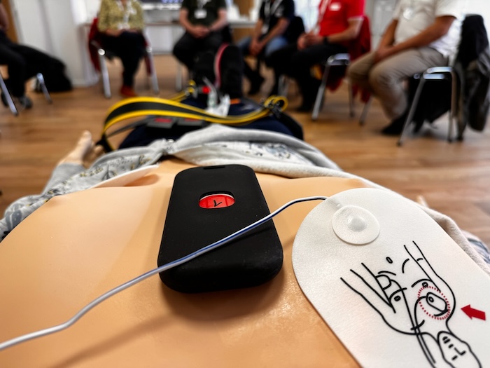 CPR Feedback und Defibrillationselektroden auf dem Oberkörper. Teilnehmer vim Hintergrund besprechen den Fall nach.
