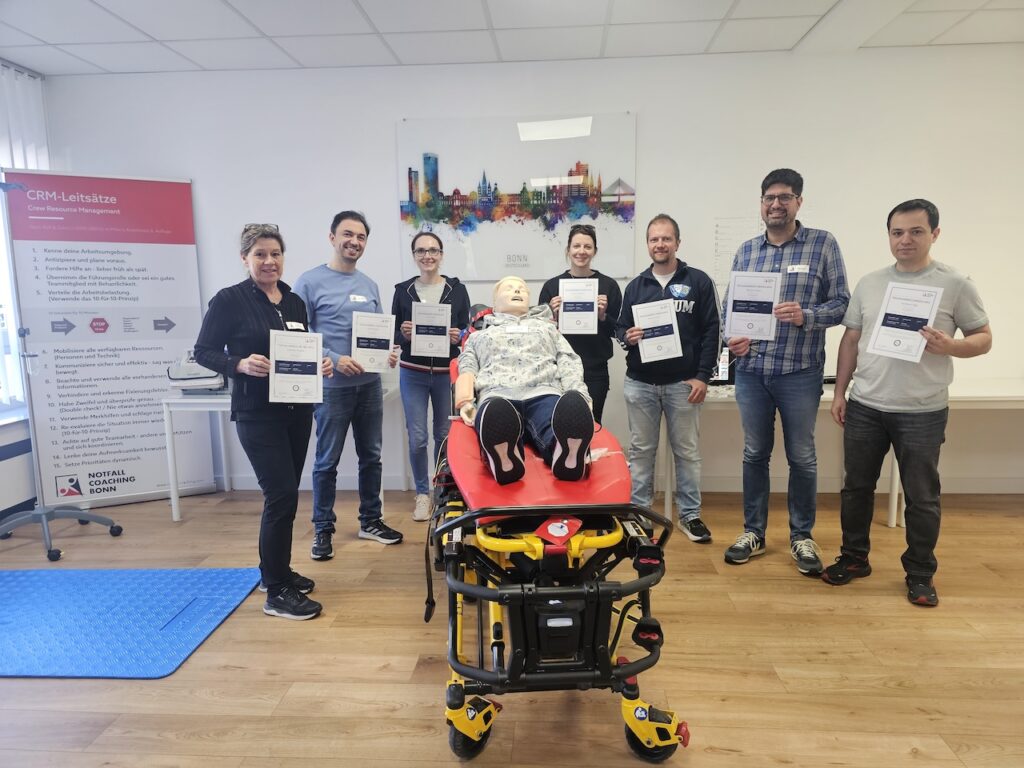 7 Personen halten ihre Teilnahmebscheinigung über den bestanden ACLS Refresher Kurs hoch. Im Vordergrund eine Simulationspuppe auf einer Rettungstrage.